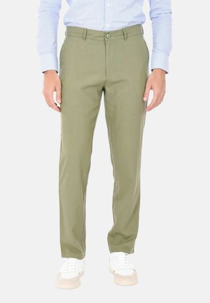 Uomo con pantaloni verde oliva, camicia a righe azzurre chiare e scarpe da ginnastica bianche, in piedi con le braccia rilassate lungo i fianchi.