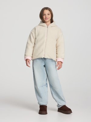 Ragazza in piedi che indossa una giacca in pile color crema con polsini rosa, jeans a gamba larga azzurri chiari e stivali marrone scuro su uno sfondo grigio semplice.