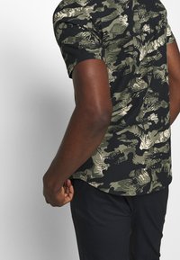 Korte mouwen shirt met een zwart-groen camouflagepatroon, textuurrijk stof en een gebogen zoom. Achteraanzicht met een relaxte pasvorm.