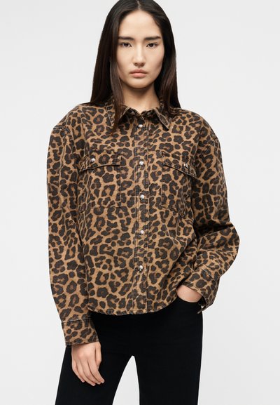 Woman wearing a brown and black leopard print buttoned jacket with flap pockets, standing with one hand in black pants pocket.