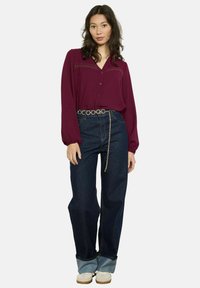 Blusa bordeaux a maniche lunghe con chiusura a bottoni e dettagli in pizzo, abbinata a jeans a gamba larga di colore blu scuro e una cintura con dettagli in catena.