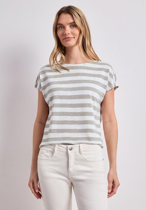 Ung kvinde med glat blondt hår iført en kortærmet grå og hvidstribet top og hvide bukser, stående foran en ensfarvet baggrund.