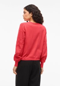 Vrouw met krullend haar, gekleed in een rood truitje met gestructureerde mouwen en een zwarte broek, staand tegen een witte achtergrond en met haar rug naar de kijker.