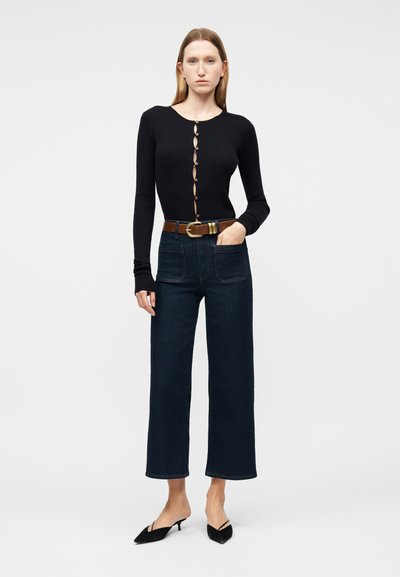 Γυναίκα που φοράει μαύρο μακρυμάνικο τοπ με κουμπιά, σκούρα ψηλόμεσα φαρδιά τζιν παντελόνια με τσέπες μπροστά, καφέ ζώνη και μαύρες μυτερές γόβες.