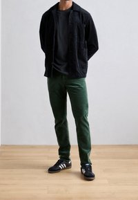 Svart manchesterjacka med två främre fickor, kombinerad med gröna manchesterbyxor. Svarta sportskor med vita ränder fullbordar outfiten.