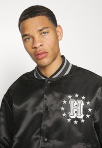 Chaqueta de béisbol de satén negro con un cuello acanalado gris, que presenta un logo blanco y estrellas en el lado izquierdo del pecho. Textura suave, cierre con botones a presión.