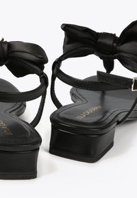Black leather sandals with a glossy finish, featuring a bow accent on the strap and a chunky heel. Textured footbed with gold logo detail.