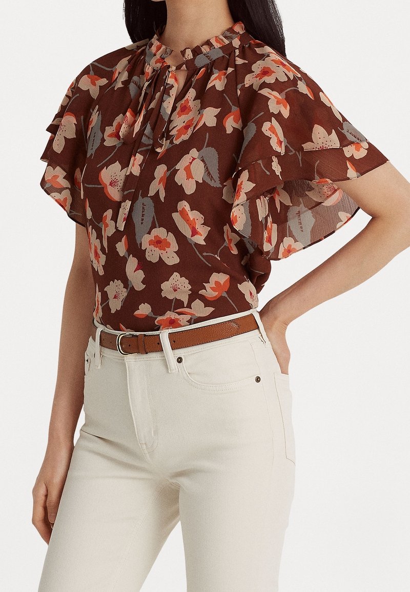 Kvinde iført brun blomstret bluse med flæsekorte ærmer og cremefarvede, højtaljede bukser med brunt bælte, hånd på hoften.