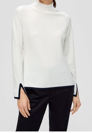 Femme portant un pull à manches longues blanc avec des bordures noires aux poignets et à l'ourlet, associé à un pantalon noir, devant un fond gris uni.