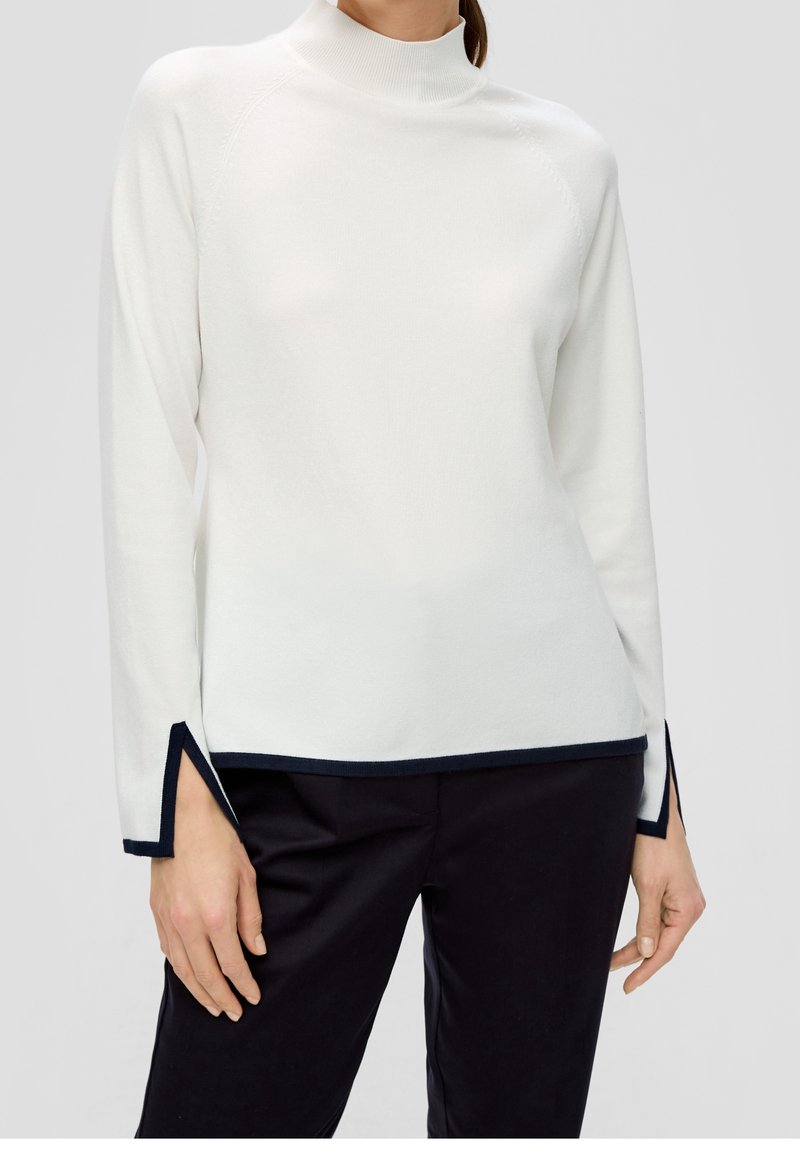 Kvinna klädd i en vit långärmad tröja med svart kant vid ärmar och nederkant, kombinerad med svarta byxor, mot en enkel grå bakgrund.