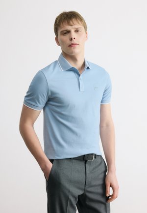 Junger Mann mit hellem Haar, der ein hellblaues Poloshirt und dunkelgraue Hose trägt, steht mit einer Hand in der Tasche vor einem schlichten Hintergrund.