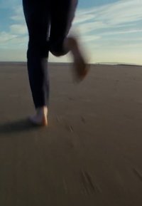 Barefoot runner on wet sand, wearing dark pants, with the shoreline and blue sky in the background. Sand texture visible beneath feet.