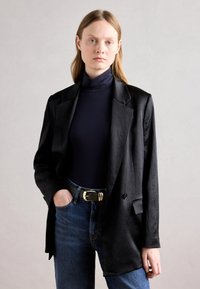Musta bleiseri, jossa on sileä pinta, ylisuurta mitoitusta ja viilipäivälliset kaulukset, yhdistettynä laivastonsiniseen poolopaitaan ja sinisiin farkkuihin. Kultainen vyönsolki näkyvissä.