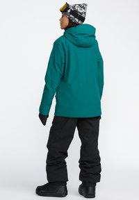 Een teal kleurige buitenjas met een capuchon, zwarte sneeuwbroek en zwarte laarzen. De jas heeft een gladde textuur en een achterzak. Geprinte beanie en goggles.