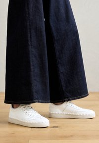 Białe sneakersy z teksturowaną gumową podeszwą i trójkątnymi akcentami. Połączone z ciemnogranatowymi spodniami w szerokim kroju. Tło z naturalnego drewnianego podłogi.