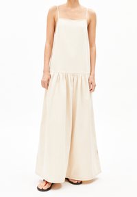 Pitkä, hihaton beige mekko, valmistettu kevyestä kankaasta, jossa on neliskanttinen pääntie, kerrostettu vyötärö ja laskeutuva helma. Minimalistinen muotoilu.