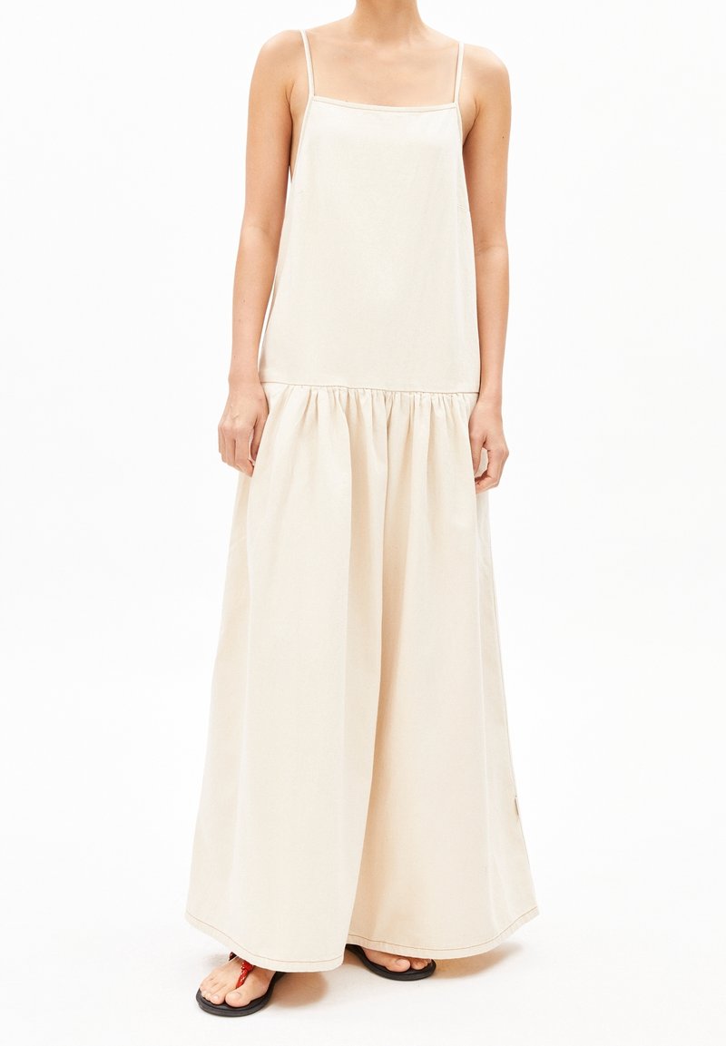 Pitkä, hihaton beige mekko, valmistettu kevyestä kankaasta, jossa on neliskanttinen pääntie, kerrostettu vyötärö ja laskeutuva helma. Minimalistinen muotoilu.