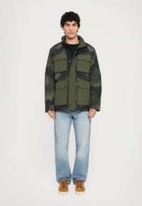 Mand i grøn og sort camouflagejakke med flere lommer, lyseblå jeans og brune sneakers, der står foran hvid baggrund.