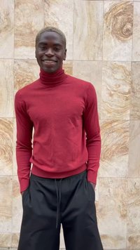 Giovane uomo sorridente con capelli biondi corti, vestito con un dolcevita rosso e pantaloni neri, in piedi contro un muro beige marmorizzato.