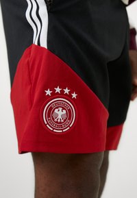 Μαύρα και κόκκινα αθλητικά σορτς με στρογγυλό κεντητό λογότυπο π�ου απεικονίζει αετούς και πέντε αστέρια, καθώς και τρεις λευκές ρίγες στο πλάι.