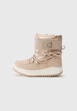 Botín impermeable beige con un acabado satinado, puño acolchado, cierre de cordones y suela de goma texturizada. Presenta un acento de logo en el costado.