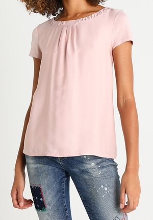 Kobieta ubrana w jasnoróżową bluzkę z krótkim rękawem i marszczonym dekoltem oraz zniszczone niebieskie dżinsy z łatami i plamami farby.