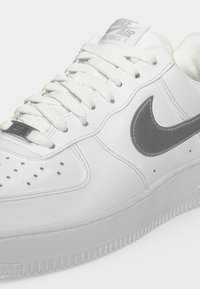 Λευκό αθλητικό παπούτσι Nike Air Force 1 με γκρι λογότυπο swoosh, λευκά κορδόνια και οπές στην περιοχή των δακτύλων, εμφανίζεται σε κοντινή λήψη.