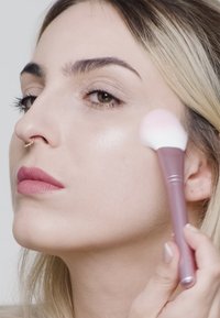 Pinceau de maquillage avec une tête de poils rose et arrondie, tenu près d'un visage en train d'appliquer un enlumineur ; teint clair, couleur de lèvres neutre, éclat subtil sur la peau.