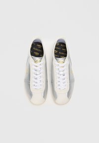 Adidași albi și argintii cu o textură netedă, având șireturi albe, un logo aurit și o talpă interioară neagră cu branding aurit.