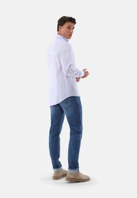 Weißes Leinenhemd mit langen Ärmeln, Kragen und abgerundetem Saum, kombiniert mit blauen Jeans und hellbraunen Stiefeln, von hinten betrachtet.