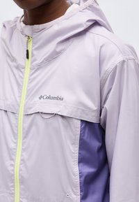 Person iført en lys lilla Columbia-jakke med hette og gul glidelås foran, som viser bryst- og skulderområdet.