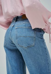 Vaqueros de mezclilla azul con un corte relajado, que presentan una cintura alta y bolsillos traseros con costuras amarillas, combinados con una blusa de color rosa claro.