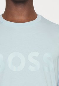 Svetle modré bavlněné tričko s kulatým výstřihem a velkým tón v tónu potiskem "BOSS" na přední straně. Měkká textura.
