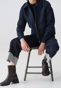 Giacca zip nera con due tasche frontali, abbinata a pantaloni in denim scuri con orlo grigio. Stivali alla caviglia marroni con punta arrotondata completano l'outfit.