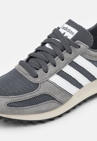 Gray sneakers featuring a mesh and suede combination, with three white stripes, round laces, and a textured black sole.