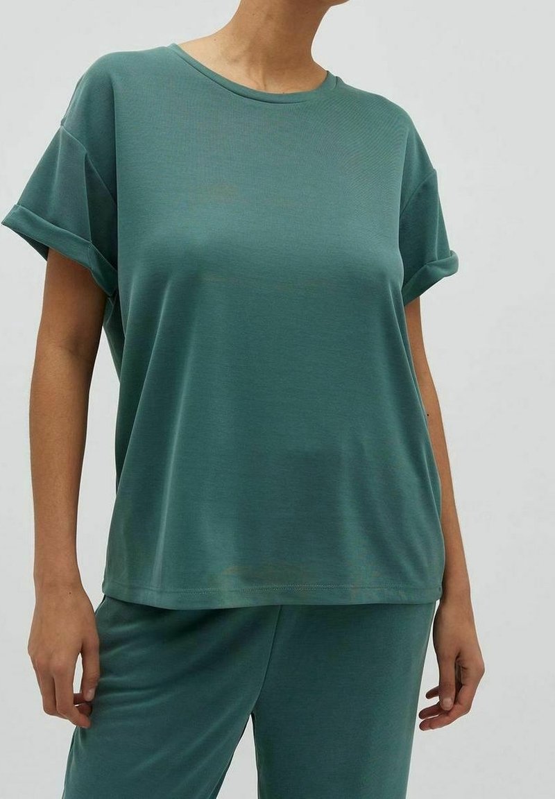 Personne portant un haut vert ample à manches courtes et un pantalon vert assorti, debout devant un fond uni.