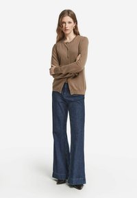 Brun cardigan i mjukt tyg med rund halsringning och knappar framtill, i kombination med högmidjade mörkdenim flarebyxor.