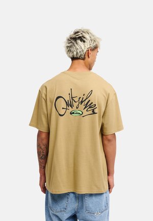 Persoon met kort blond haar die een beige Quiksilver t-shirt en blauwe spijkerbroek draagt, gezien van achteren tegen een effen achtergrond.
