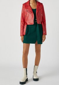 Femme portant une veste en cuir rouge courte, une robe en maille verte ceinturée avec une fente devant, et des bottines blanches avec semelles noires et côtés élastiques.