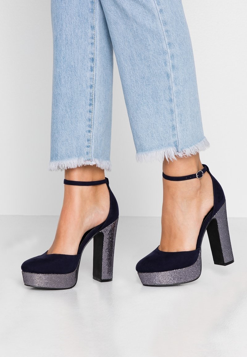 Chaussures à talons hauts en daim bleu marine avec une plateforme métallique argentée et un talon carré. Présente une bride au niveau de la cheville et un design à bout rond.
