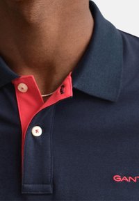 Gros plan d'une personne portant un polo bleu marine avec une patte intérieure rouge et des boutons blancs, montrant une partie du logo GANT.