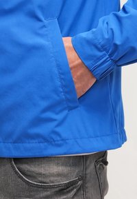 Blå, lätt jacka med elastiska ärmslut, sid ficka och mjuk textur. Har en rak skärning och snygg sömnad.