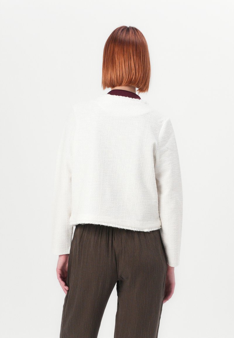 Witte, textured cropped jas met lange mouwen en rafelige zoom, gedragen over bruine, geplooide broek; gezien van de achterkant.