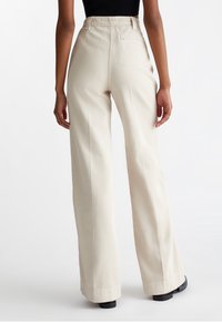 Højtaljede, vide beige bukser lavet af bomuldsblanding. Har sidelommer og en baglommer. Glat tekstur med pressede folder.