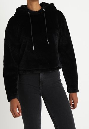 Schwarzer Kapuzenpullover mit flauschiger Oberfläche und Kordelzug, getragen mit schwarzen, hochtaillierten, eng anliegenden Jeans an einer stehenden Person.