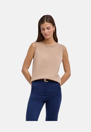 Femme debout, mains derrière le dos, portant un haut sans manches beige, un pantalon bleu marine, un collier en or et des boucles d'oreilles anneaux, sur un fond uni.