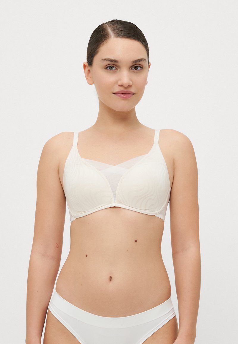 Light beige bra with a wavy textured pattern, molded cups, and adjustable straps. Features a sheer mesh panel at the front.