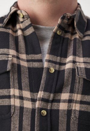 Beige-schwarz kariertes Hemd mit Knöpfen über einem grauen T-Shirt mit Rundhalsausschnitt, Ausschnitt und oberer Brustbereich sichtbar.