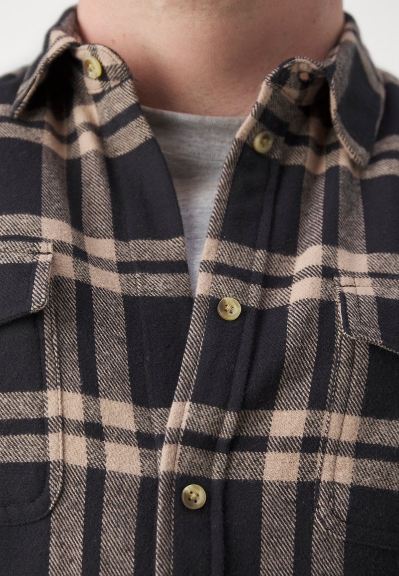 Chemise à carreaux beige et noire boutonnée par-dessus un t-shirt gris à col rond, montrant le cou et la partie supérieure de la poitrine.