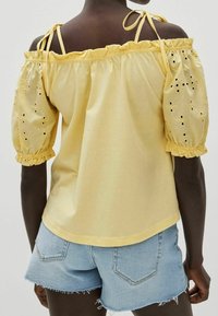 Gul off-the-shoulder top med puffærmer, med hulsyning, samlet elastik ved halsen og manchetterne, parret med denimshorts.